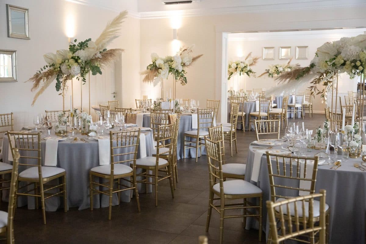 Festsaal mit runden Tischen in Grau, goldenen Chiavari-Stühlen und hohen Blumendeko-Ständern aus Pampasgras und weißen Rosen