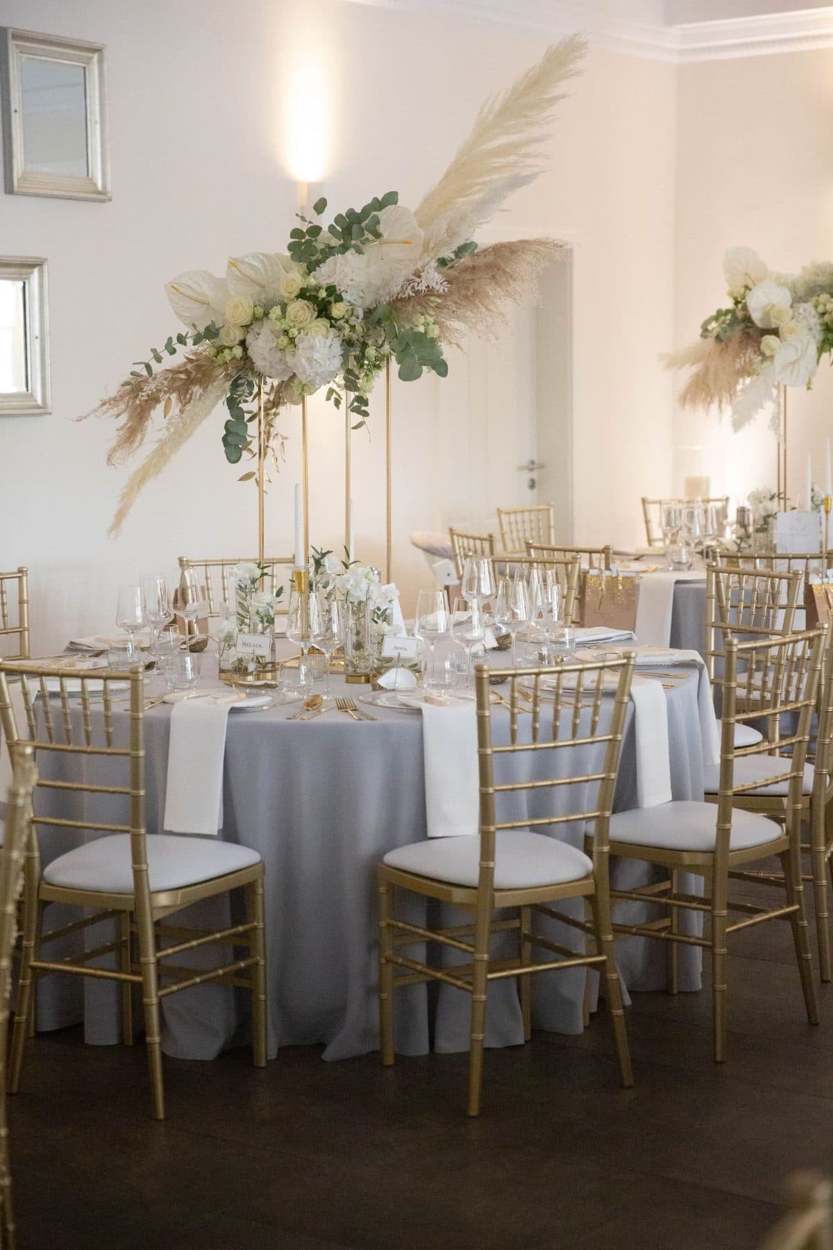 Runder Hochzeitstisch mit hoher Blumendeko aus weißen Rosen, Pampasgras und Eukalyptus auf goldenem Ständer, grauer Tischdecke und goldenen Chiavari-Stühlen