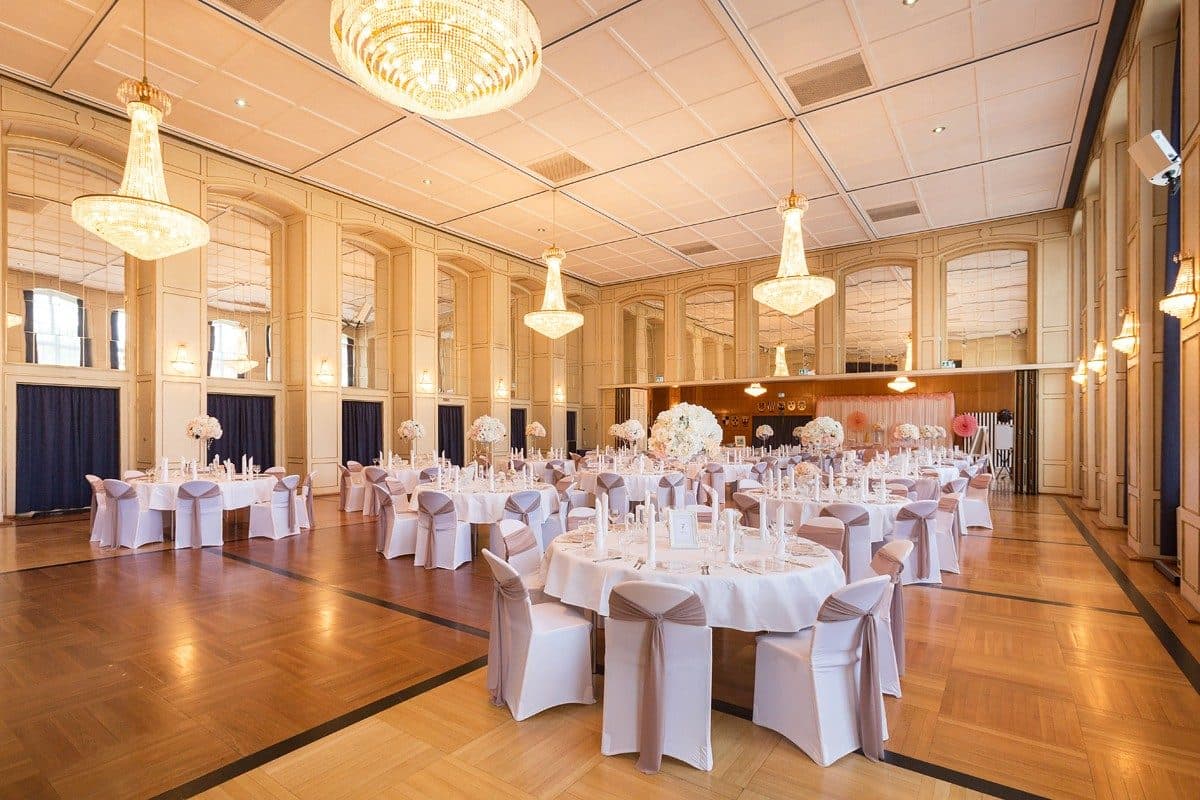 Festsaal mit Kristall-Kronleuchtern, Spiegelwänden, Parkettboden und runden Hochzeitstischen mit taupefarbenen Stuhlschleifen
