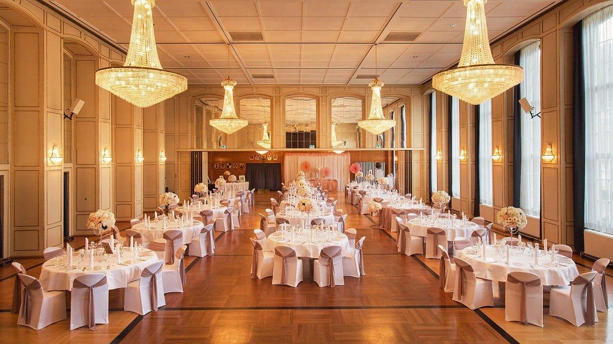 Festsaal mit Kristall-Kronleuchtern, Spiegelwänden und runden Hochzeitstischen mit taupefarbenen Stuhlschleifen, Blick zur Bühne