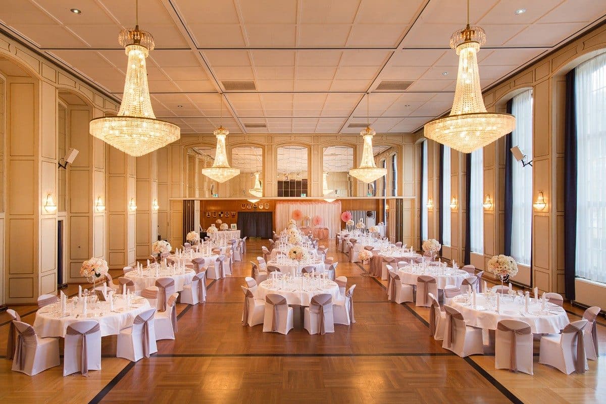 Festsaal mit Kristall-Kronleuchtern, Spiegelwänden und runden Hochzeitstischen mit taupefarbenen Stuhlschleifen