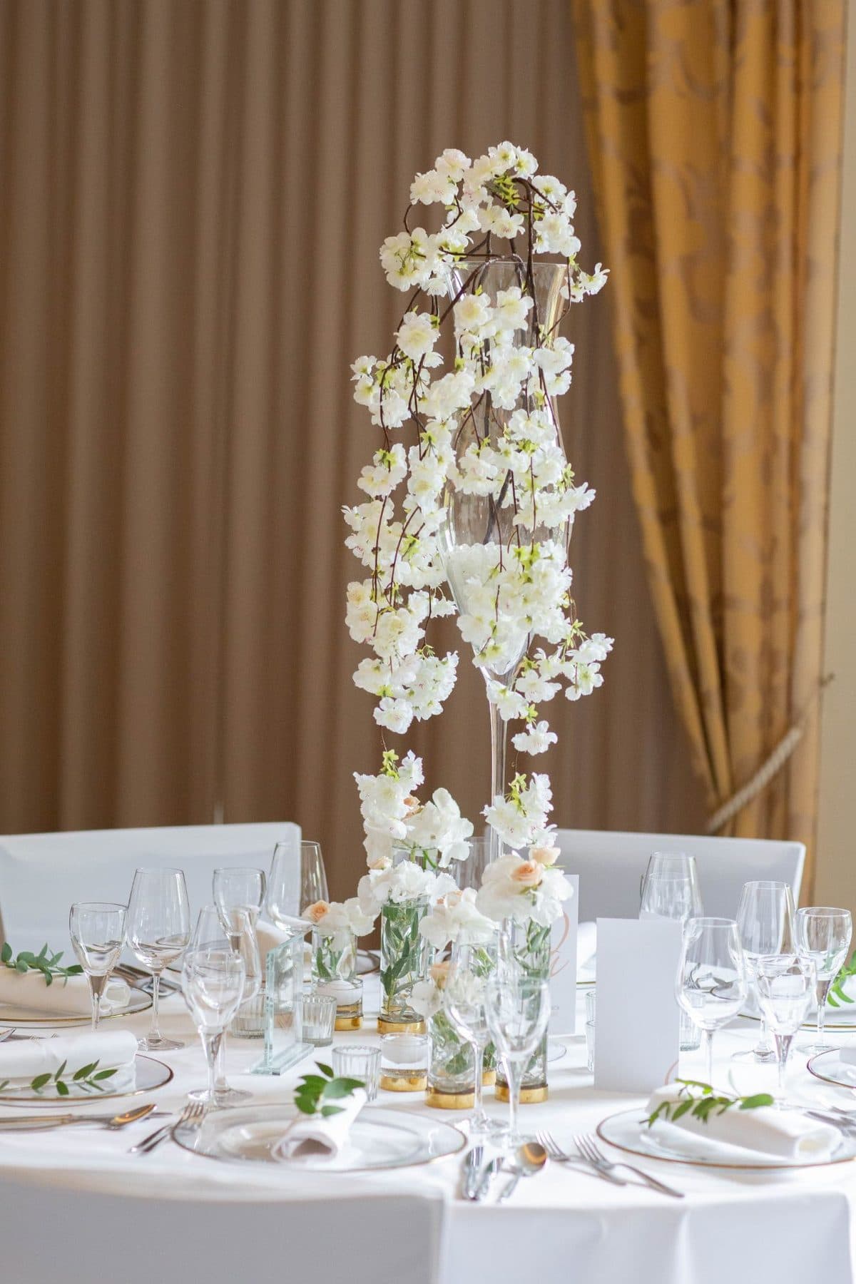 Hohe schlanke Glasvase mit weißem Kirschblütenzweig auf Hochzeitstisch mit Tischnummer und Kristallglas-Details