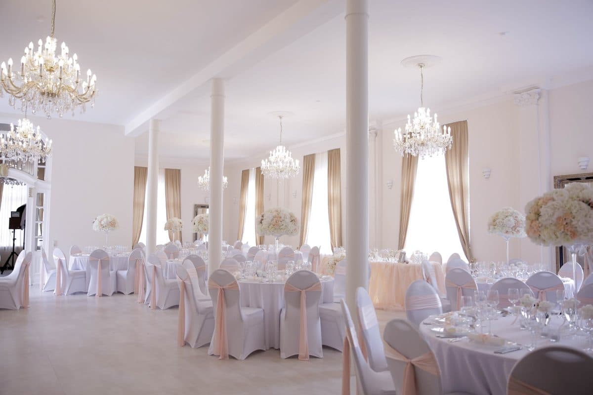 Festsaal im Alten Kurhotel Pforzheim mit Kristall-Kronleuchtern, weißen Säulen, goldenen Vorhängen und runden Hochzeitstischen mit rosé Stuhlschleifen