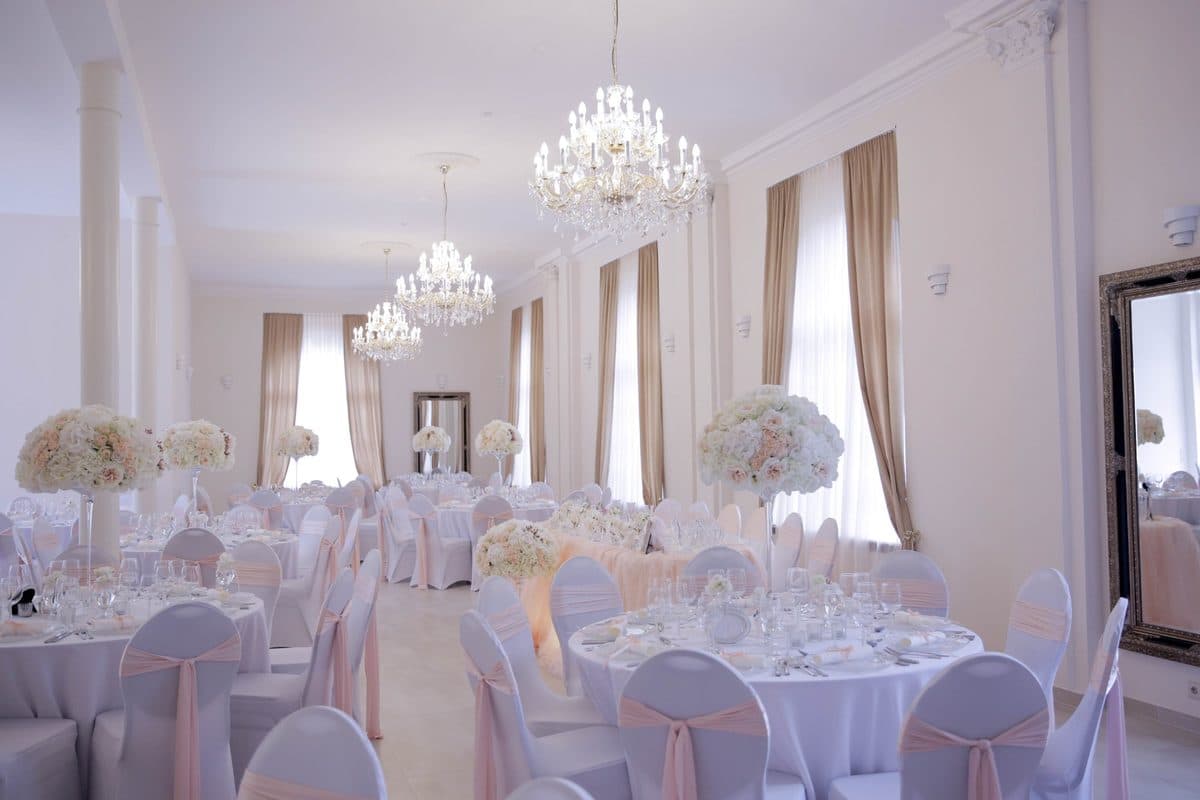 Festsaal im Alten Kurhotel Pforzheim mit Kristall-Kronleuchtern, weißen Säulen und runden Hochzeitstischen mit rosé Stuhlschleifen