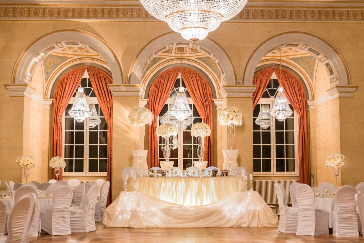 Brauttisch im König Ludwig I.-Saal vor Rundbogenfenstern mit Kristallkronleuchtern und hohen weißen Blumengestecken