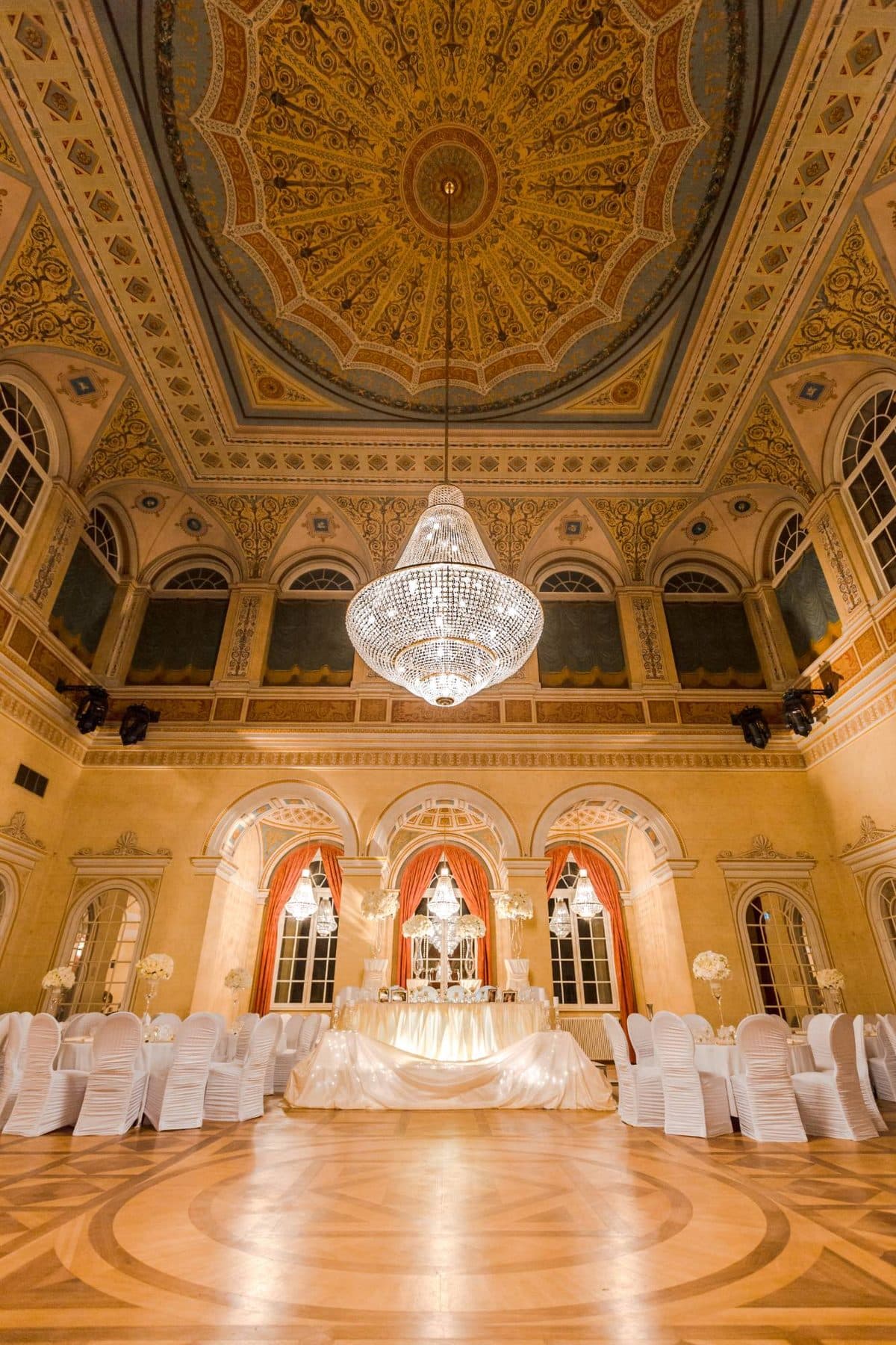 König Ludwig I.-Saal in Bad Brückenau mit prunkvollem Deckenfresko, Kristallkronleuchter und weiß gedeckten Hochzeitstischen