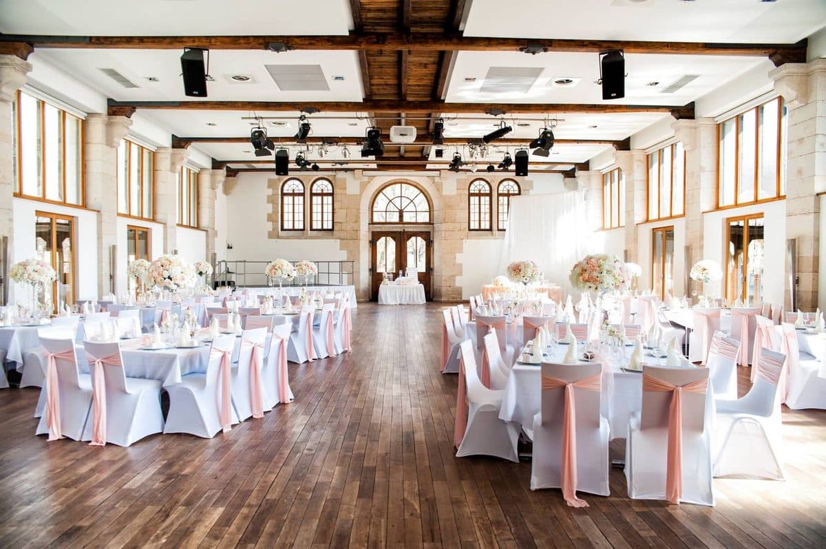 Hammerschmiede Königsbronn bei Tageslicht mit hohen Rundbogenfenstern und runden Hochzeitstischen mit rosé Stuhlschleifen