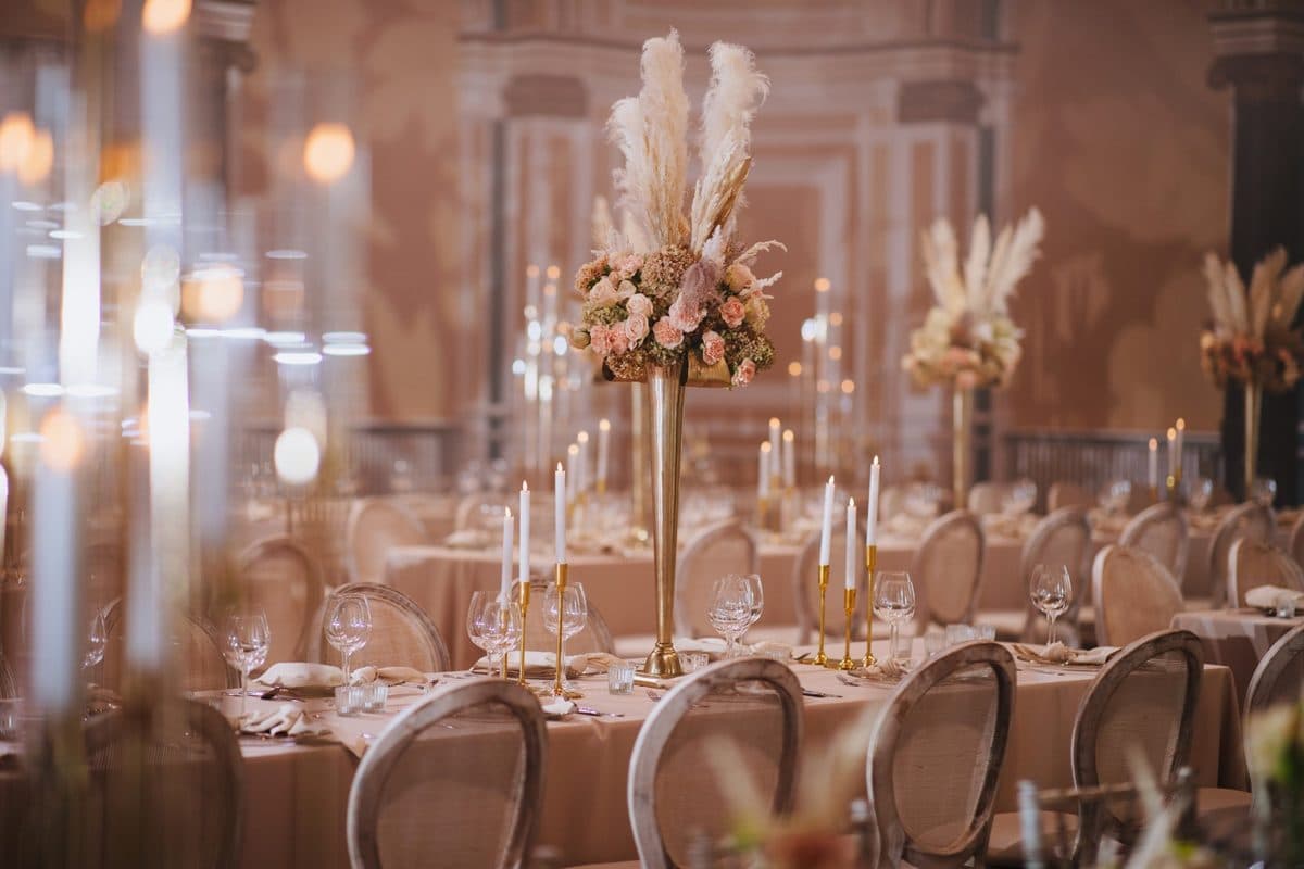 Festsaal mit hohen goldenen Trompetenvasen, Pampasgras und pastellfarbenen Blumen