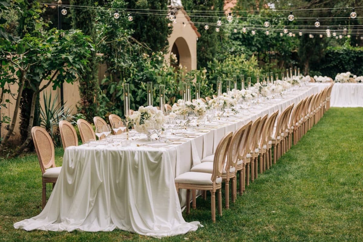 Lange Gartentafel mit weißer Tischdecke, Blumen und Lichterkette im mediterranen Garten