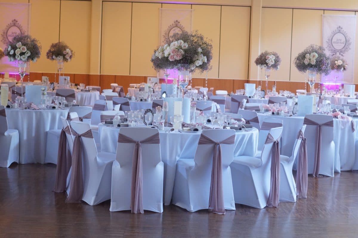 Festsaal mit weißen Stuhlhussen, mauvefarbenen Bändern und hohen Blumengestecken aus Rosen und Schleierkraut