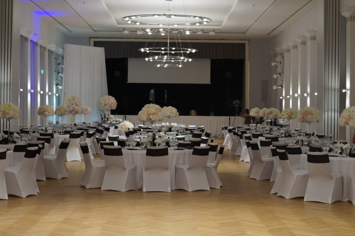 Festsaal mit weißen Stuhlhussen, schwarzen Spitzenbändern und hohen Blumengestecken vor der Bühne