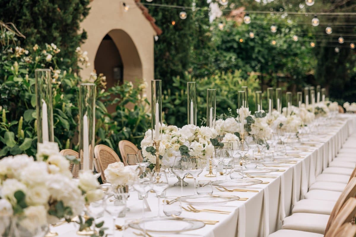 Lange Gartentafel im Grünen mit goldenen Kerzenständern, weißen Rosen und Lichterketten