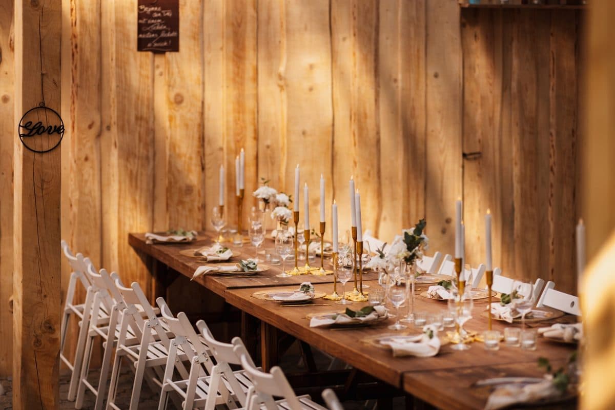 Lange festlich gedeckte Hochzeitstafel unter Holzpavillon mit goldenen Kerzenleuchtern, Glasteller und Kreidetafel