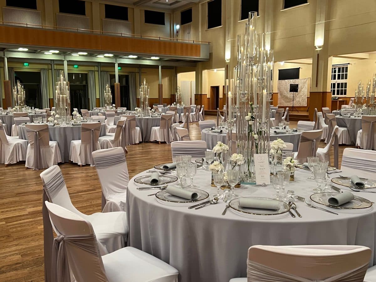 Runde Hochzeitstische mit hohen Kristall-Kerzensäulen, silbernen Stuhlhussen und Platztellern in der Stadthalle Haslach