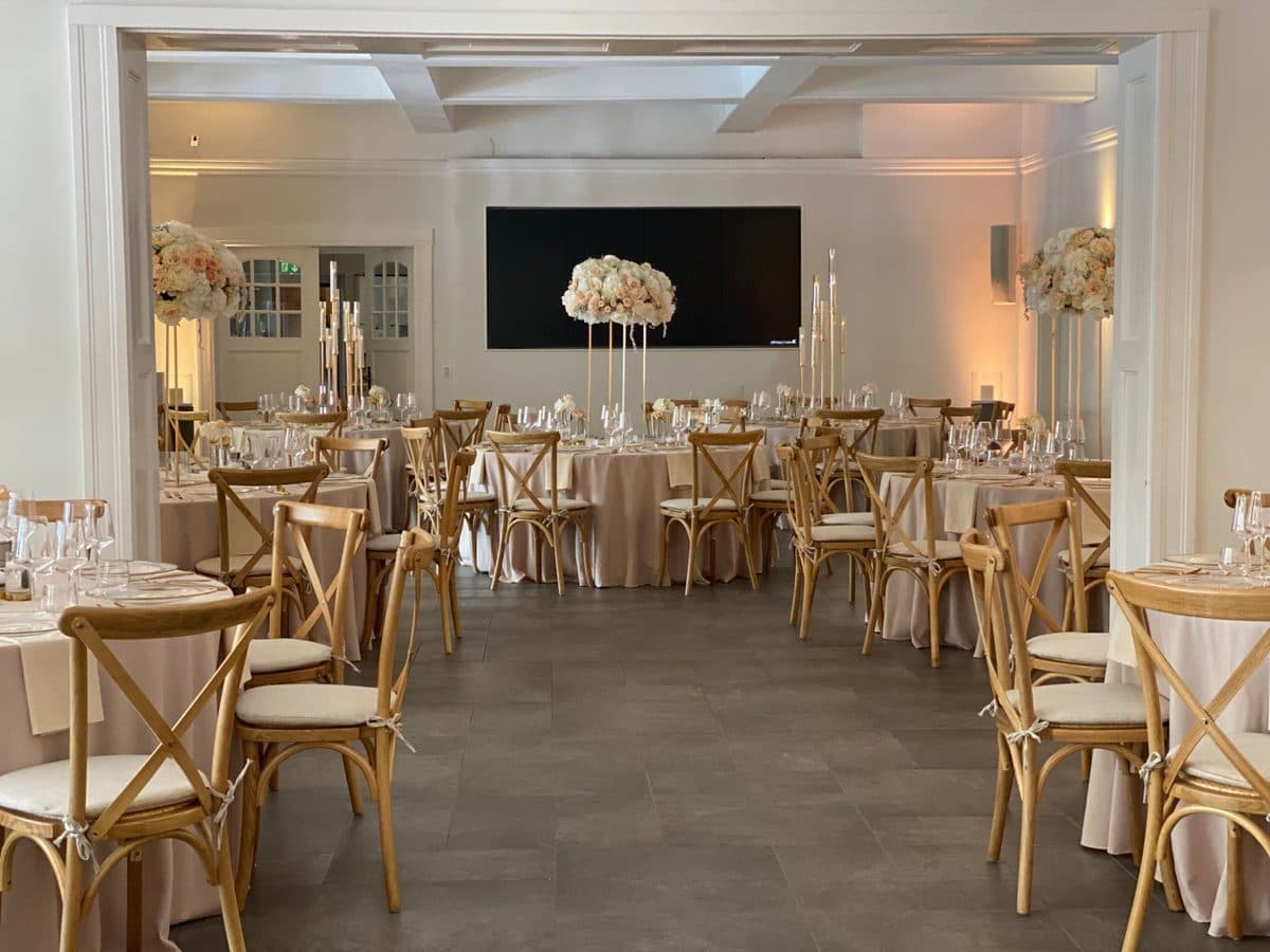 Festsaal mit runden Tischen in Taupe, hellen Holzstühlen und hohen weiß-rosé Blumendeko-Ständern